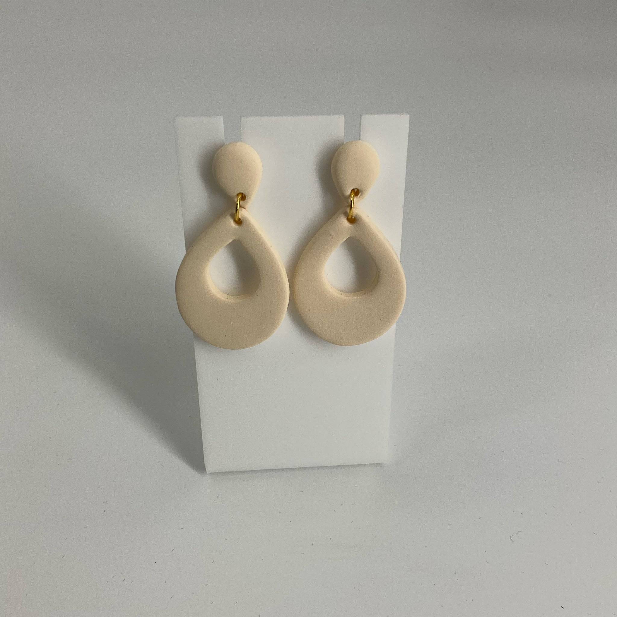 Handmade cream-colored teardrop dangle earrings with gold-tone connectors, displayed on a white stand. Stylish accessory for modern outfits.
