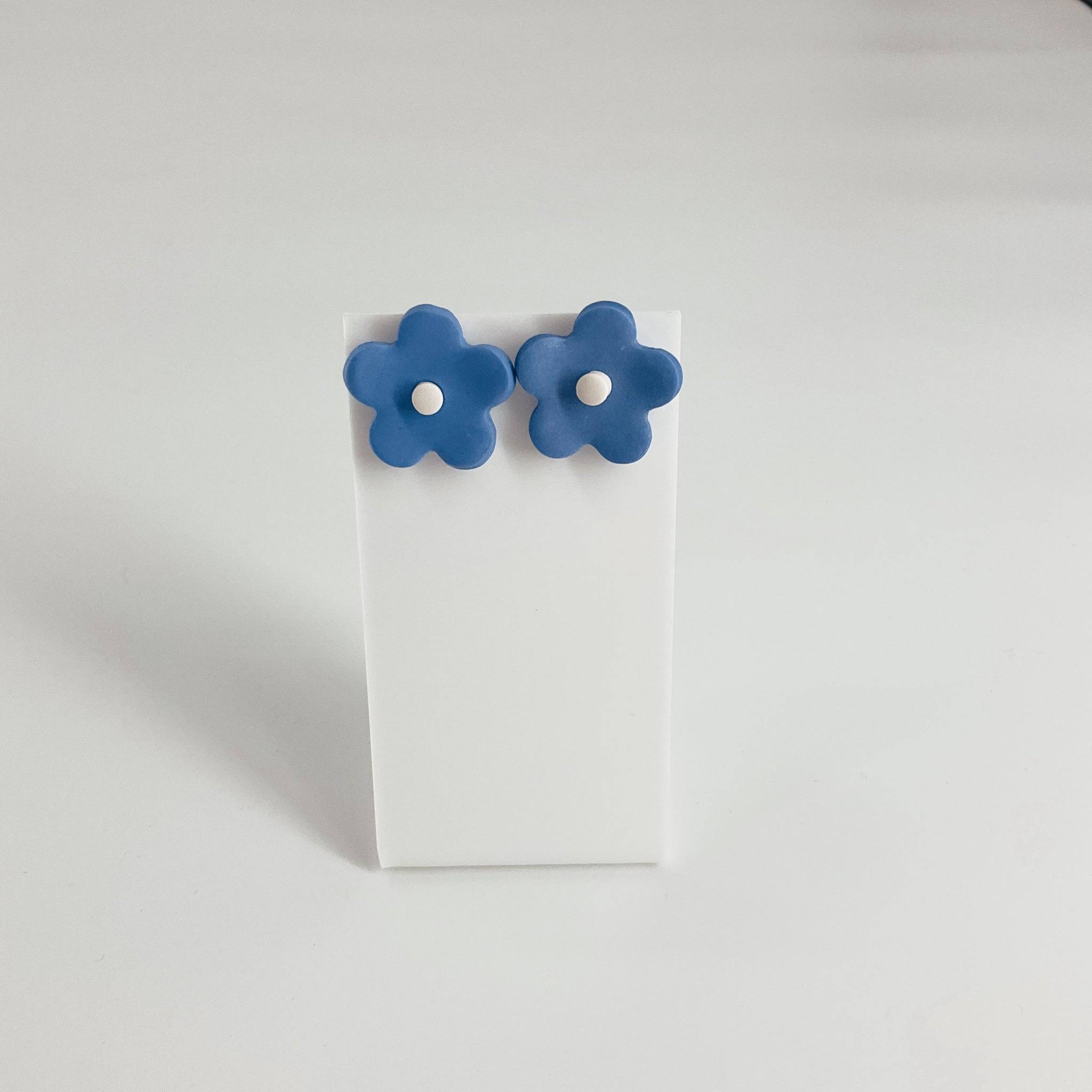 Handmade blue flower earrings with a minimalist design, featuring five-petal shapes and white centers, displayed on a white stand.